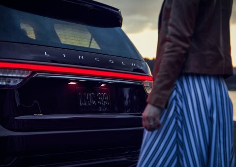A person is shown near the rear of a 2024 Lincoln Aviator® SUV as the Lincoln Embrace illuminates the rear lights | Brinson Lincoln of Corsicana in Corsicana TX